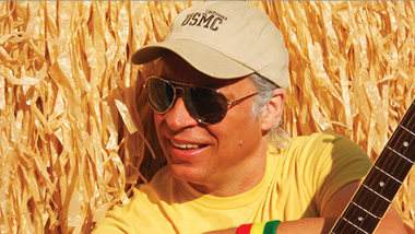 Photo of Jimmy Bluffett holding guitar laying in corn
