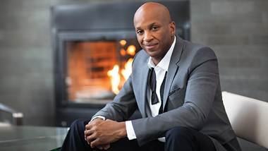 Donnie McClurkin sitting on a couch in front of a fireplace.