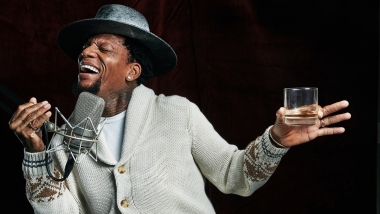 DL Hughley singing while holding a glass of bourbon with black background
