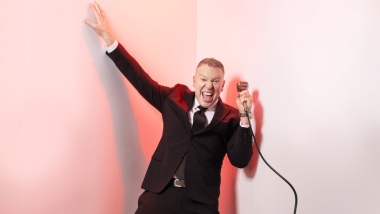 Gary Owen in a suit, holding a mic with pink background