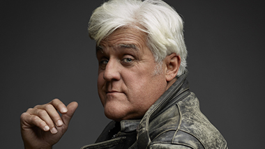 Close up of Jay Leno looking into the camera on a dark gray background.